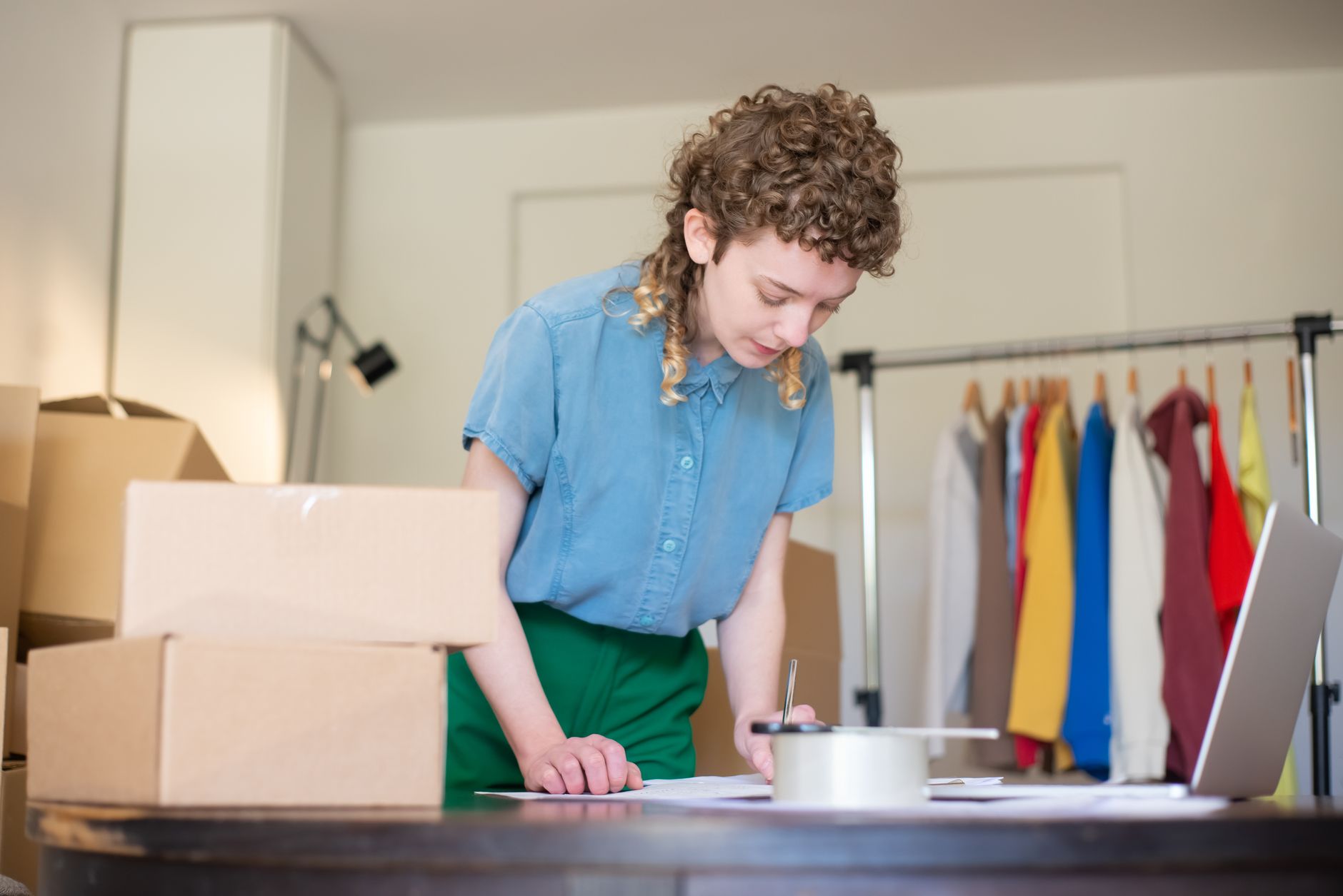Femme préparant des commandes chez elle, entourée de cartons et d'un portant à vêtements, illustrant une activité indépendante gérée à domicile.
