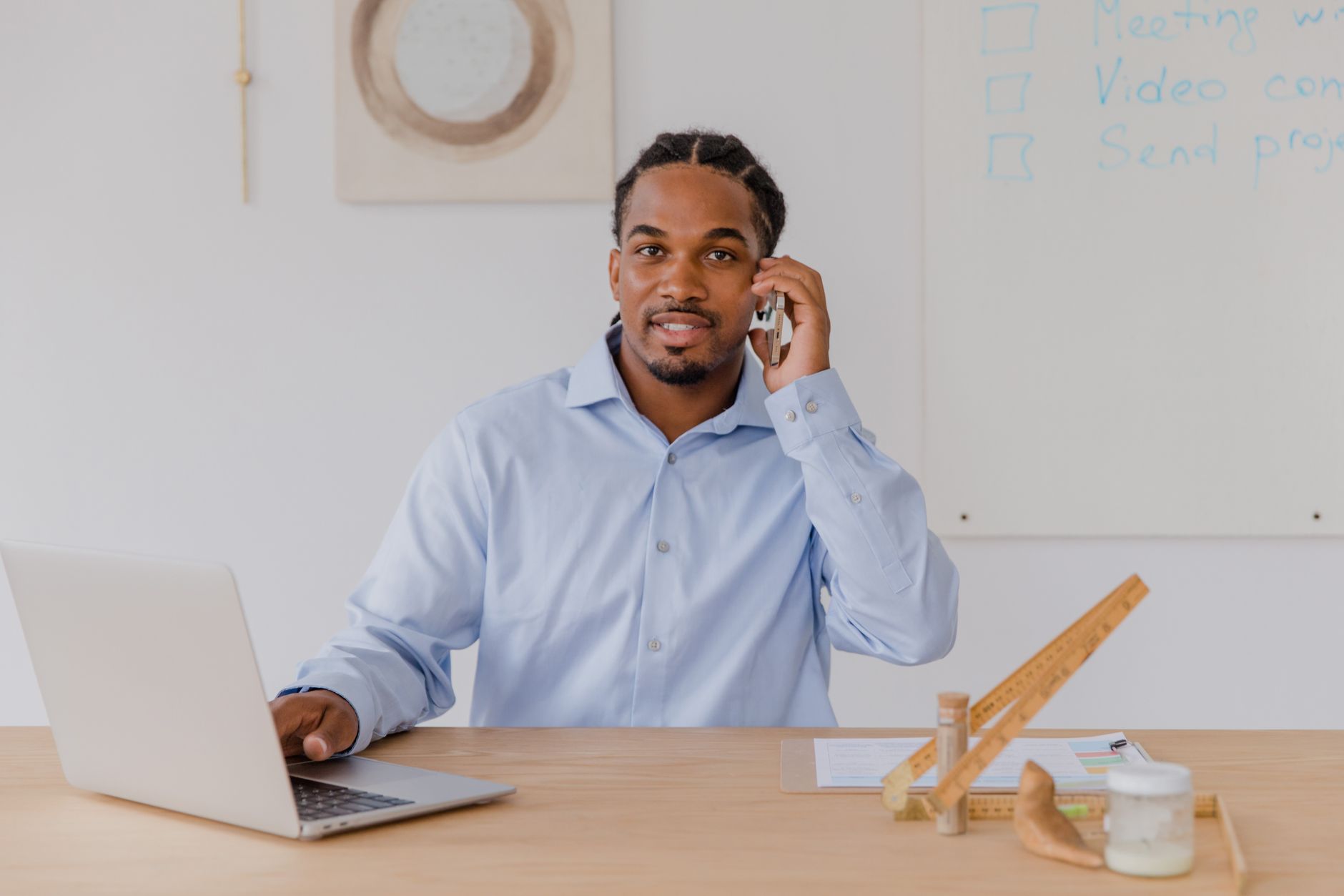 Jeune homme souriant à son bureau, smartphone en main et ordinateur portable ouvert, dans un environnement de travail professionnel.